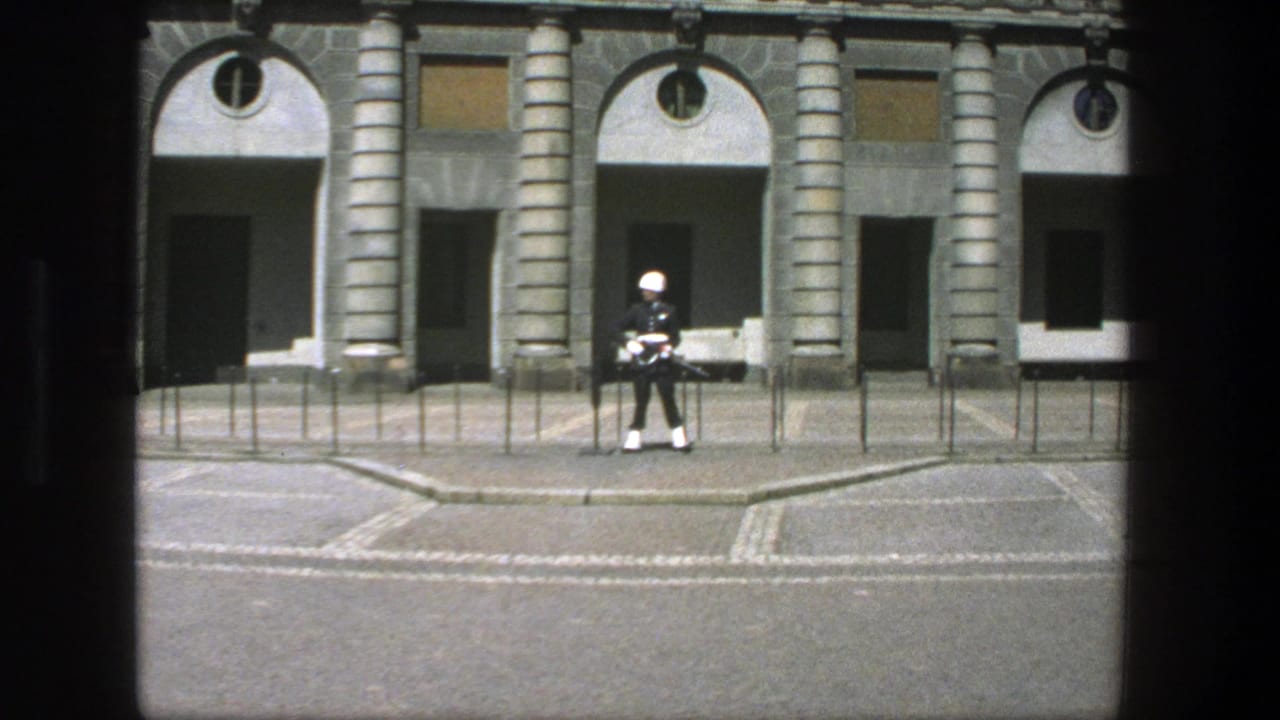 Guard in dress uniform stands near the center of a paved frontage with arched doors behind.