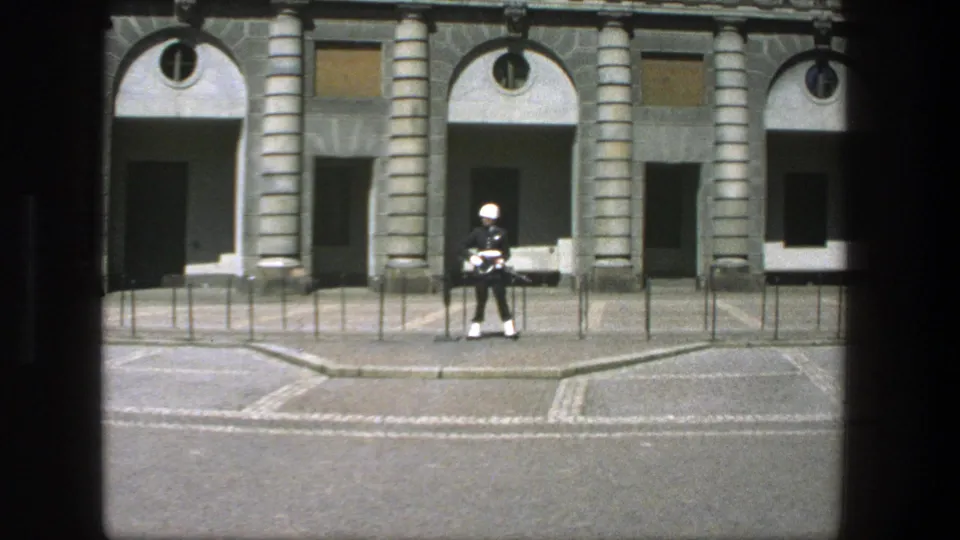 1975 Stockholm frame with a dress-uniformed guard holding a rifle before a stone facade with arched entrances.