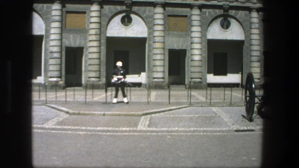 1975 Stockholm 8mm frame showing a military guard and the wheel of a cannon near a formal stone building.