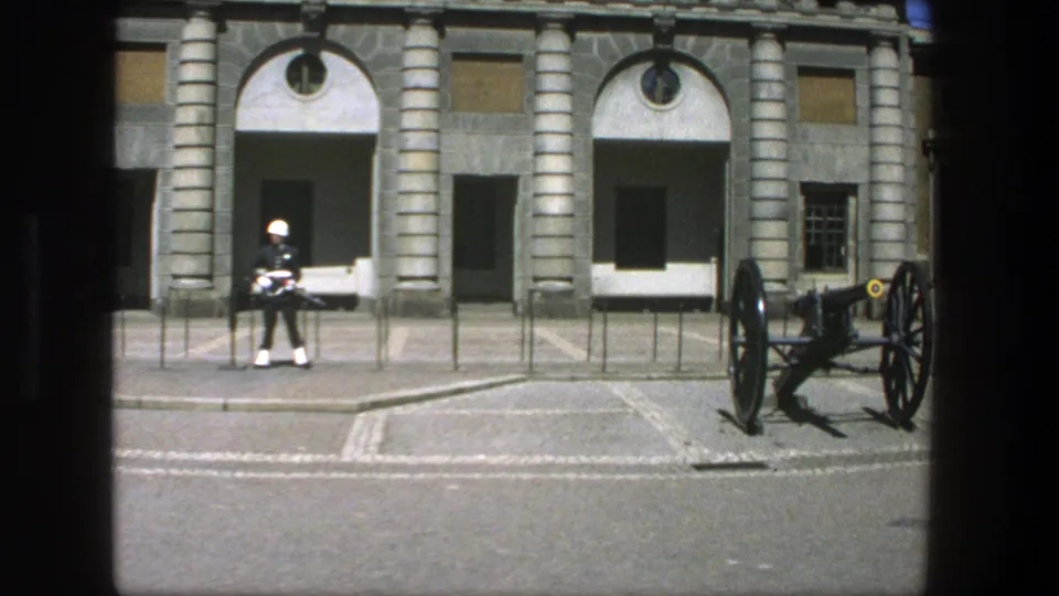 1975 Stockholm frame with a uniformed military guard and cannon outside a grand stone building.