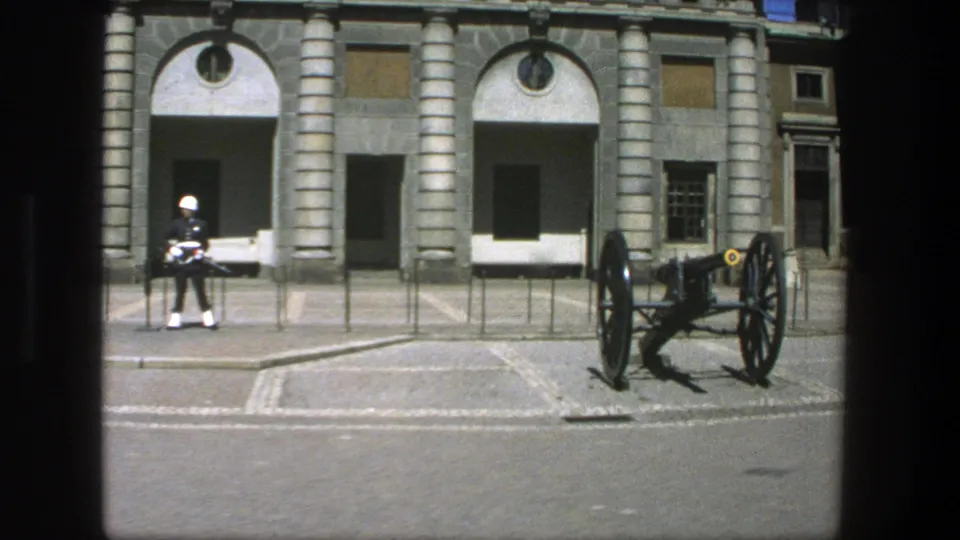 1975 Stockholm 8mm frame showing a white-helmeted guard near a displayed cannon in front of a formal stone facade.