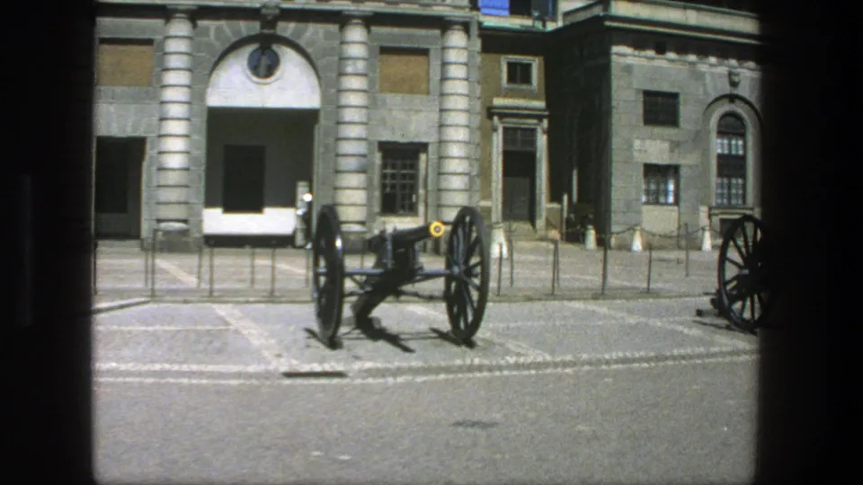 1975 Stockholm 8mm frame showing a large cannon in the foreground and a guard near the stone building behind it.