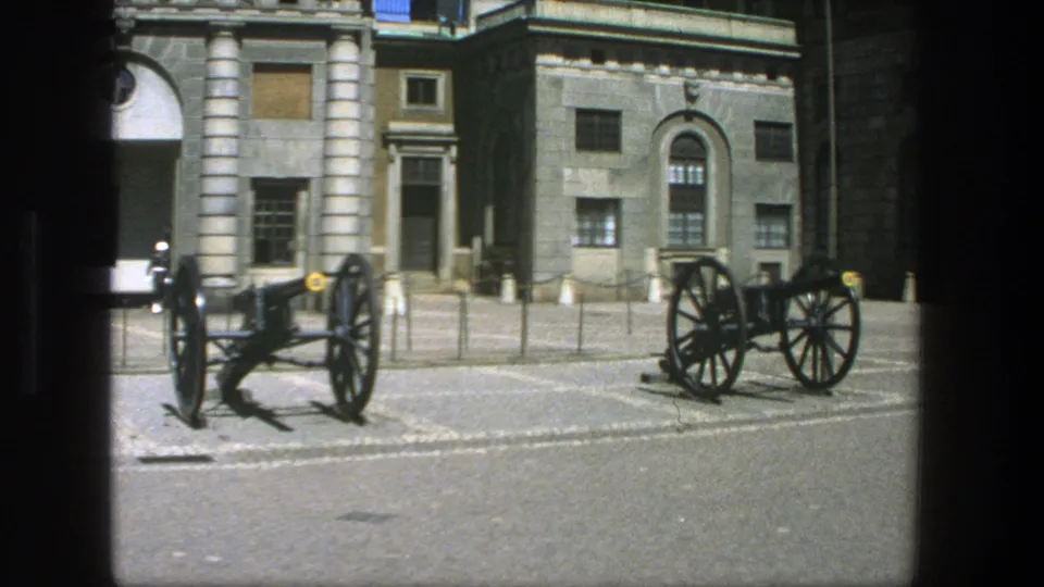 1975 Stockholm 8mm frame showing a paved courtyard, two displayed cannons and a stone building with columns and arched windows.