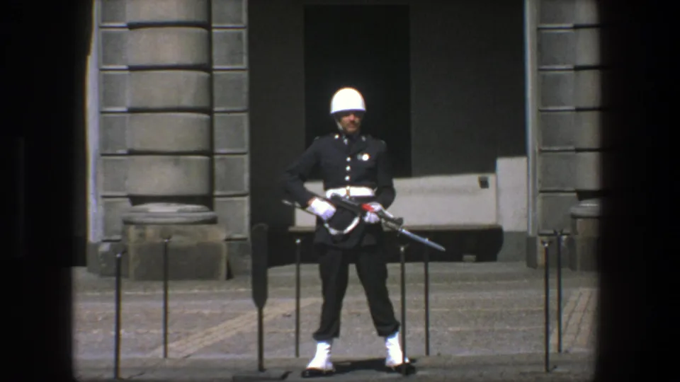 1975 Stockholm frame of a ceremonial armed guard in dark uniform, white helmet and gloves standing at attention by a stone facade.