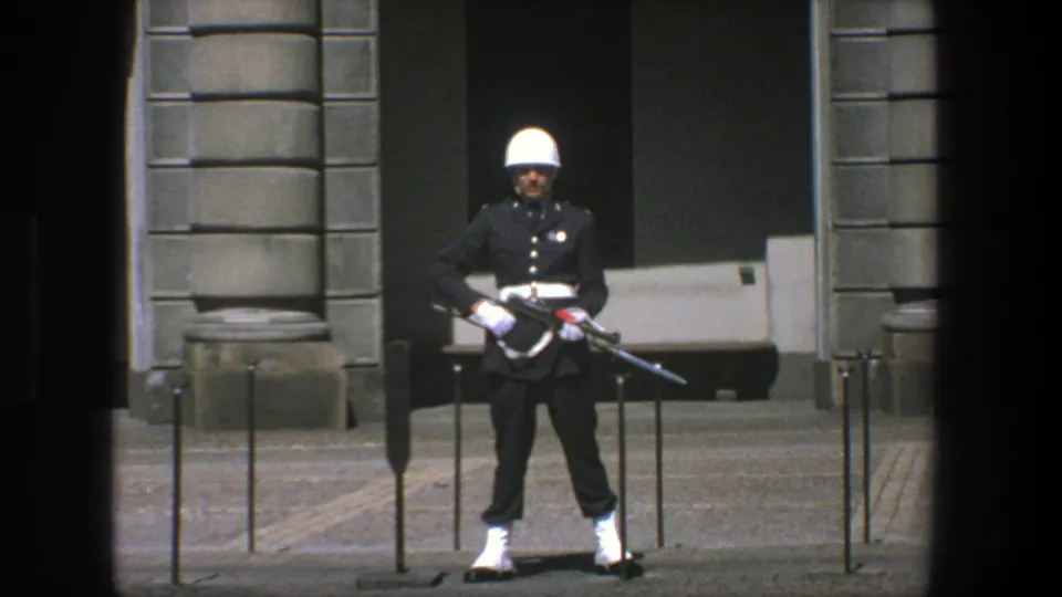 A Stockholm ceremonial guard stands with rifle and white helmet against a gray stone building in the 1975 8mm footage.