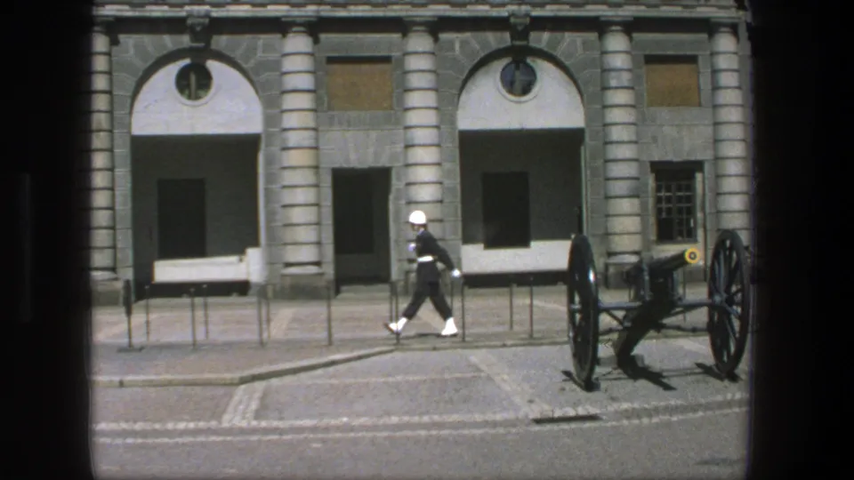 A ceremonial guard marches across a Stockholm courtyard with a cannon, arched entrances, and stone columns visible behind him.