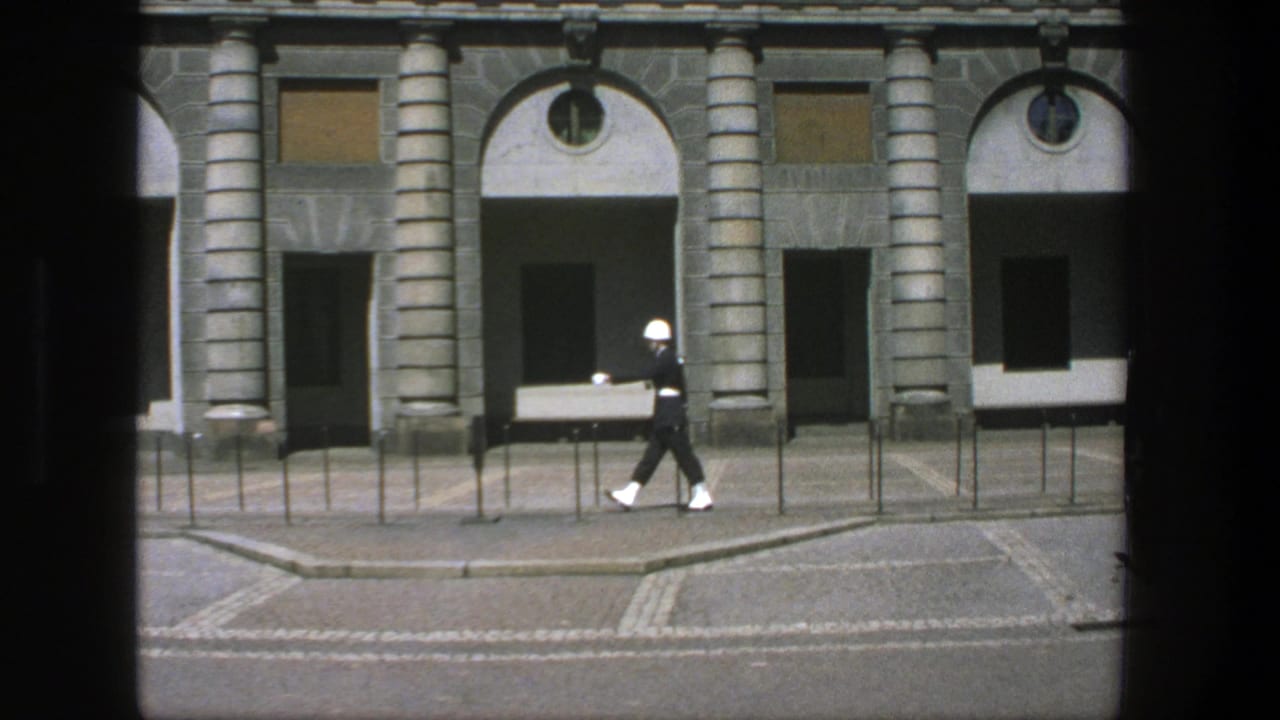 Uniformed guard marching past a stone building with arched entrances.