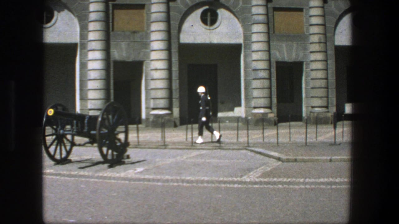 In Stockholm, Sweden in 1975, a white-uniformed guard marches near a historic cannon before a formal stone building.