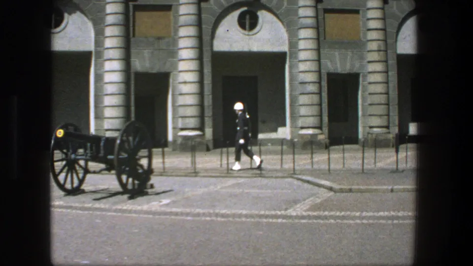 In Stockholm, Sweden in 1975, a white-uniformed guard marches near a historic cannon before a formal stone building.
