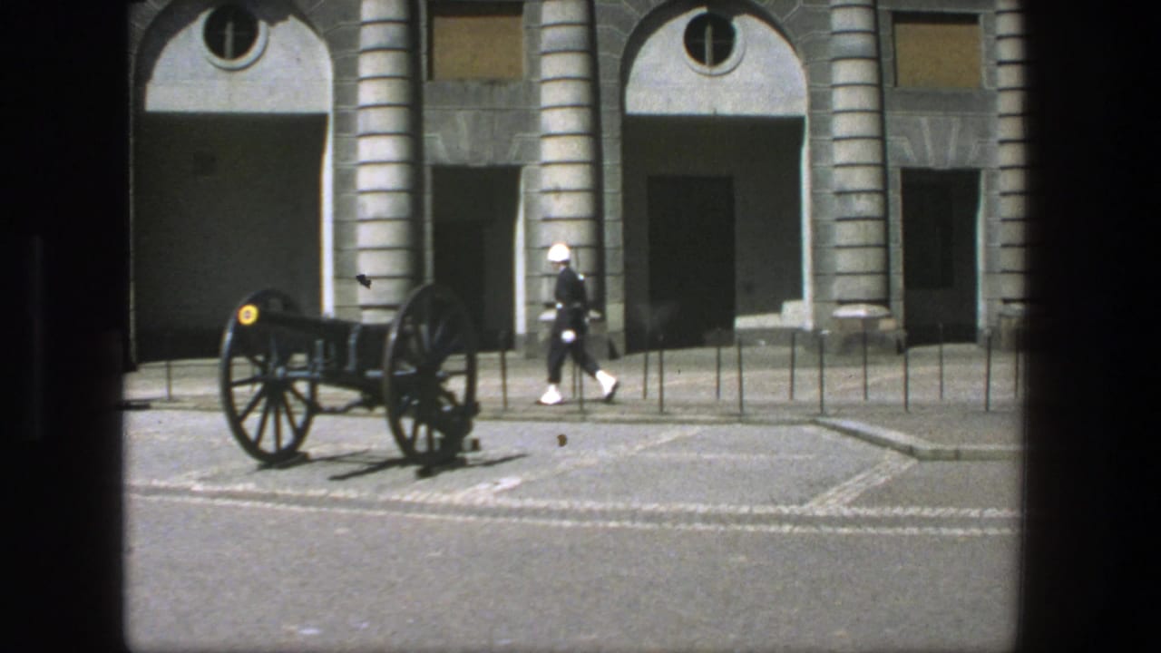 A white-uniformed guard continues across the courtyard near a cannon and arched building facade in 1975 Stockholm.