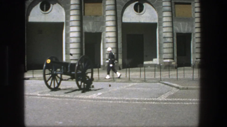 A white-uniformed guard continues across the courtyard near a cannon and arched building facade in 1975 Stockholm.