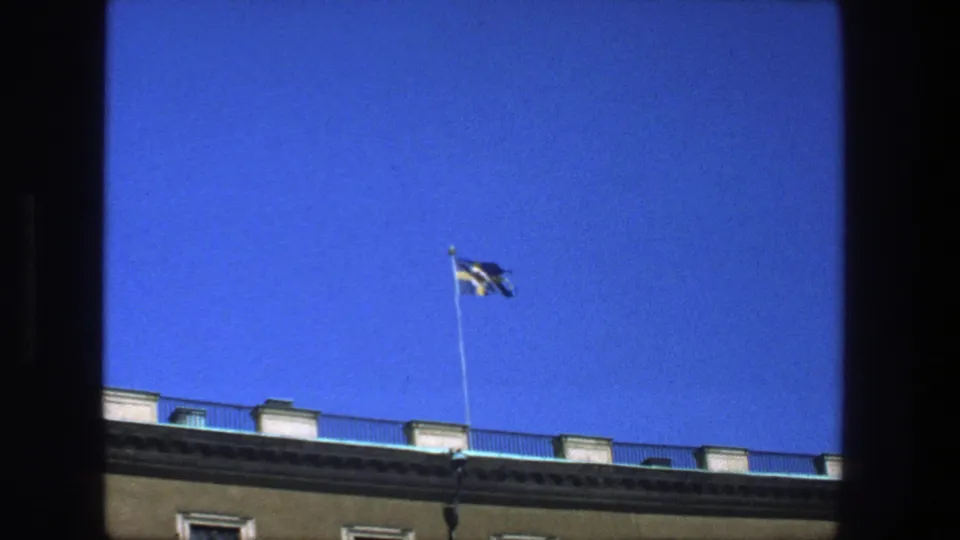 A Swedish flag flies over a building roofline against clear blue sky in the Stockholm home-movie footage.