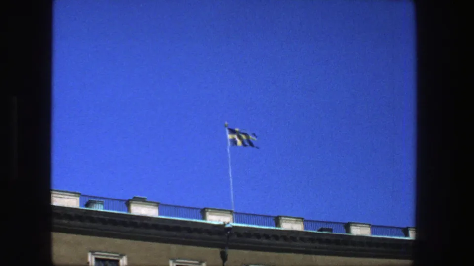 The Swedish flag remains visible above the roofline, framed by blue sky in the 1975 Stockholm scene.