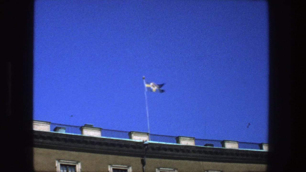 Stockholm home-movie frame with the Swedish flag fluttering above a grand stone building and clear blue sky.