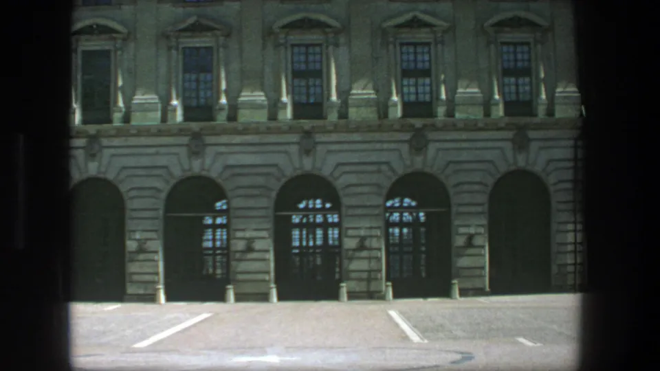 1975 Stockholm 8mm frame showing the lower facade of a formal stone building, dark arches, window grids, and a quiet paved forecourt.