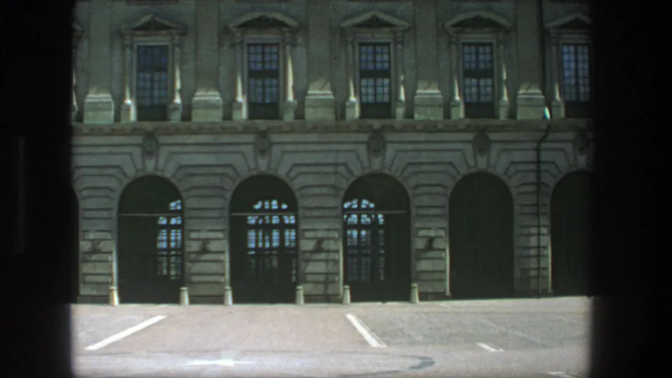 1975 Stockholm home-movie still of a grand stone exterior with arched entrances, tall windows, bollards, and a dark foreground frame.