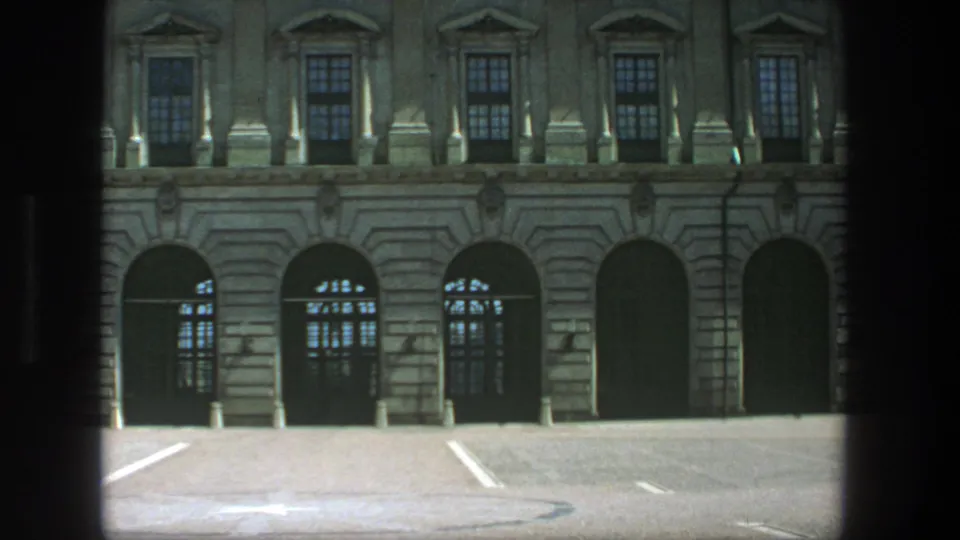 1975 8mm view in Stockholm of a tan classical building facade with arched openings and an empty paved lot in front.