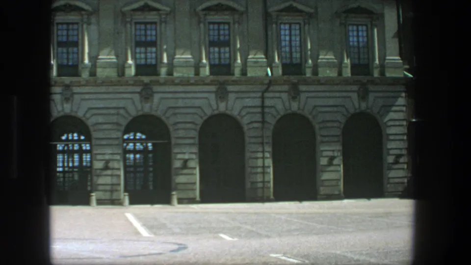 Vintage 8mm Stockholm view of a tan classical building and quiet parking area under bright daylight.