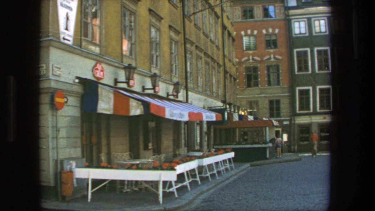 1975 Stockholm street view with empty outdoor cafe tables, a striped awning, cobblestones, signs, and tall building facades.
