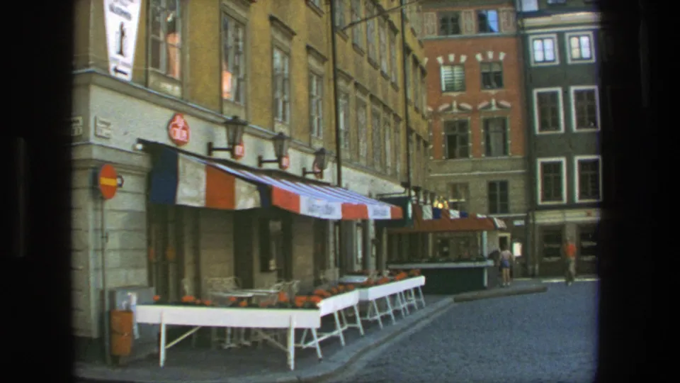 1975 Stockholm street view with empty outdoor cafe tables, a striped awning, cobblestones, signs, and tall building facades.