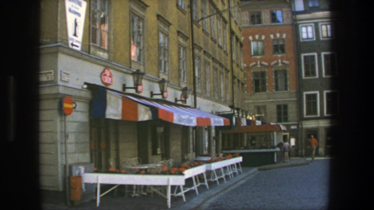 1975 Stockholm cafe frontage with white outdoor tables, empty chairs, striped awning, cobblestones, and pedestrians farther down the street.