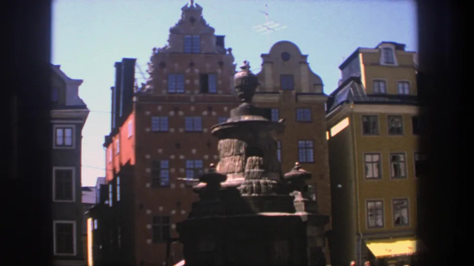 1975 Stockholm view of an ornate fountain near tall historic facades, with bright sky and street-level shopfronts partly visible.