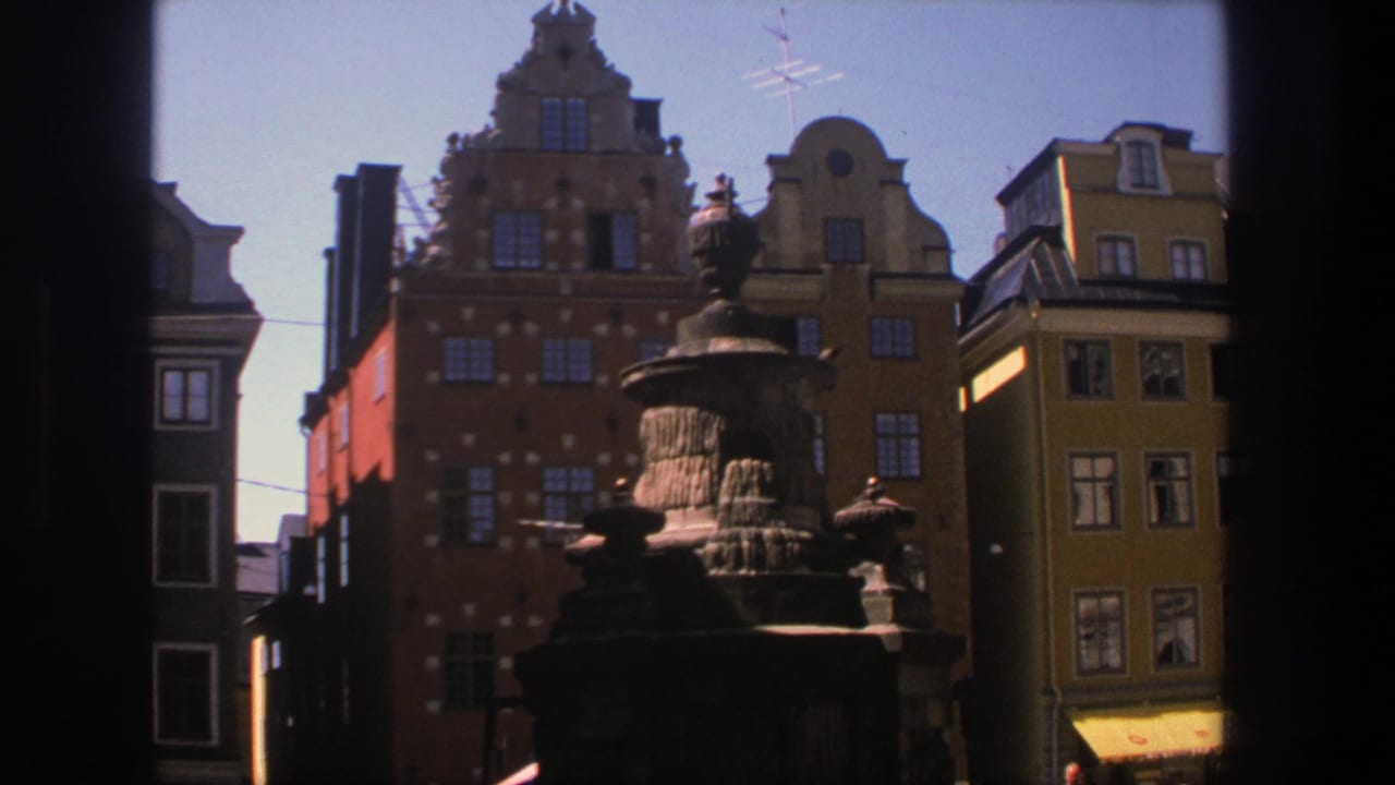 1975 8mm Stockholm view of a stone fountain and ornate colorful buildings seen through dark framing edges.