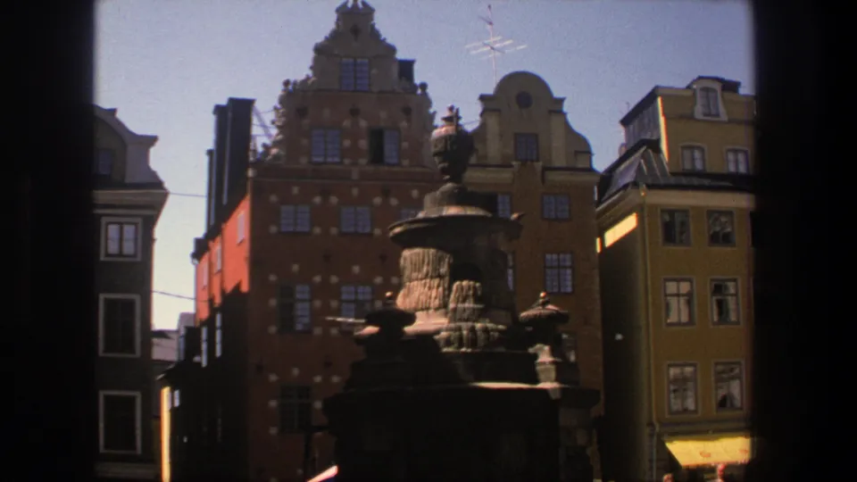 1975 Stockholm home-movie frame showing a shadowed fountain against sunlit historic building fronts.