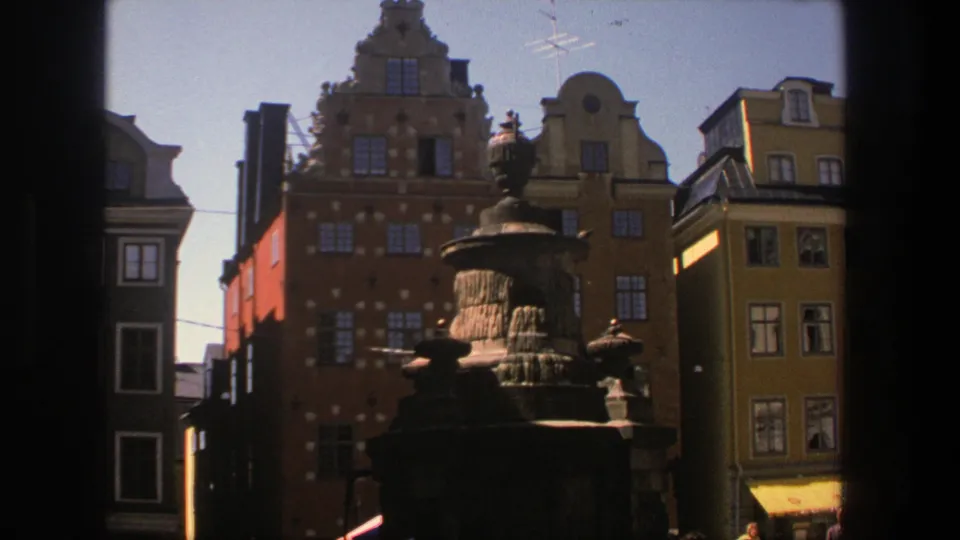 8mm Stockholm frame with a stone fountain, colorful historic facades, and a small yellow awning in daylight.