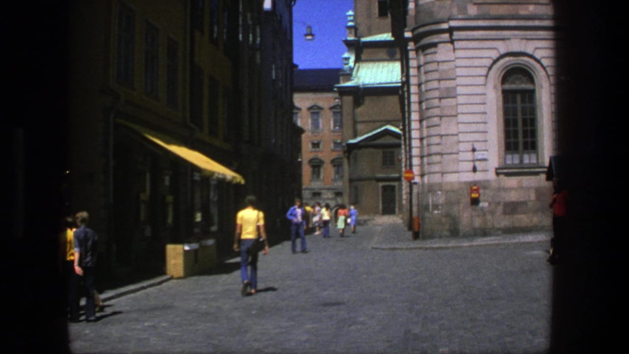 Pedestrians walk through a sunny cobblestone alley beside a stone church wall and shaded storefronts.