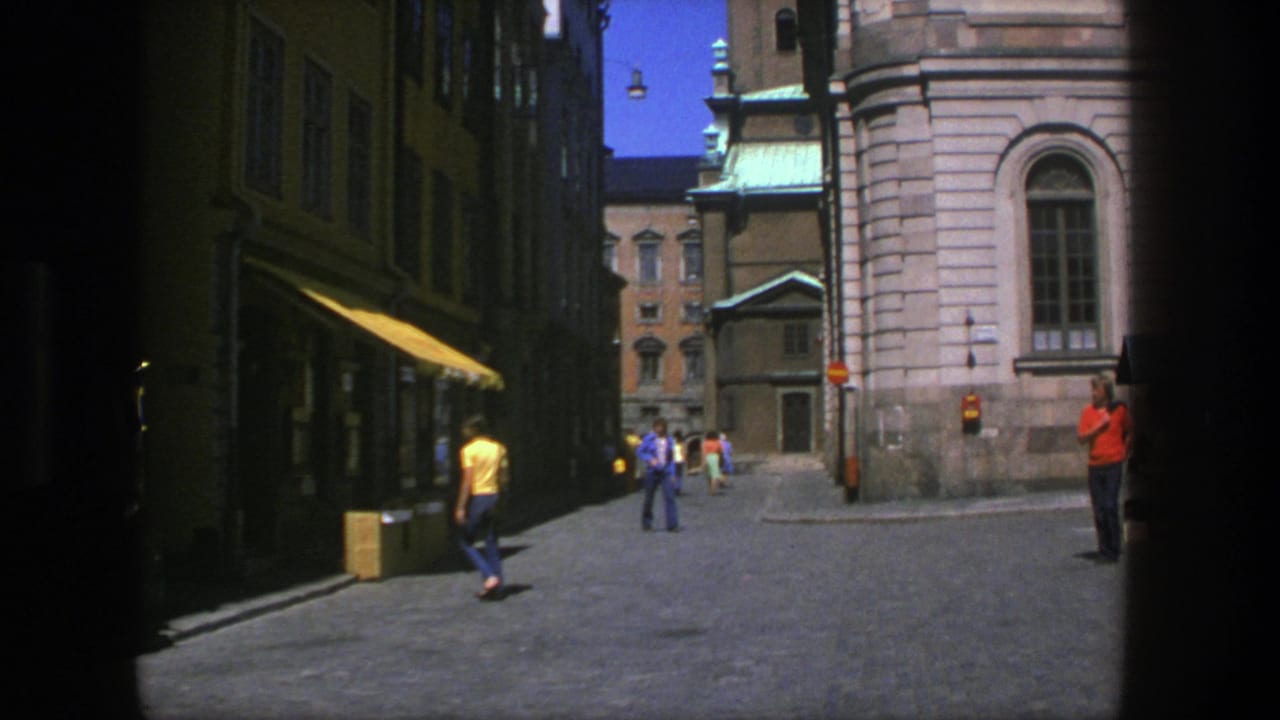 1975 Stockholm alley scene with a pedestrian in yellow, distant walkers, shaded shopfronts, and a large stone church wall.