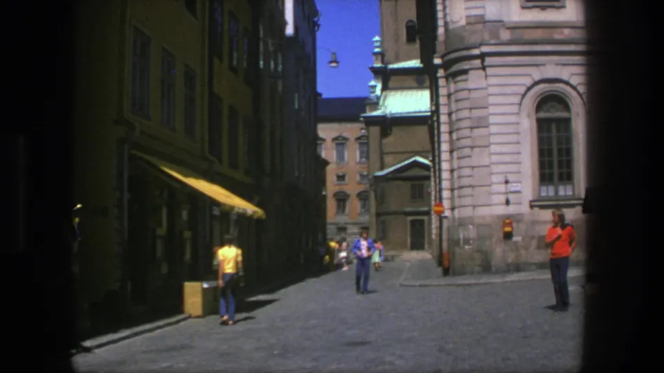 1975 Stockholm street footage showing pedestrians in colorful clothing near a shaded storefront and historic stone church exterior.