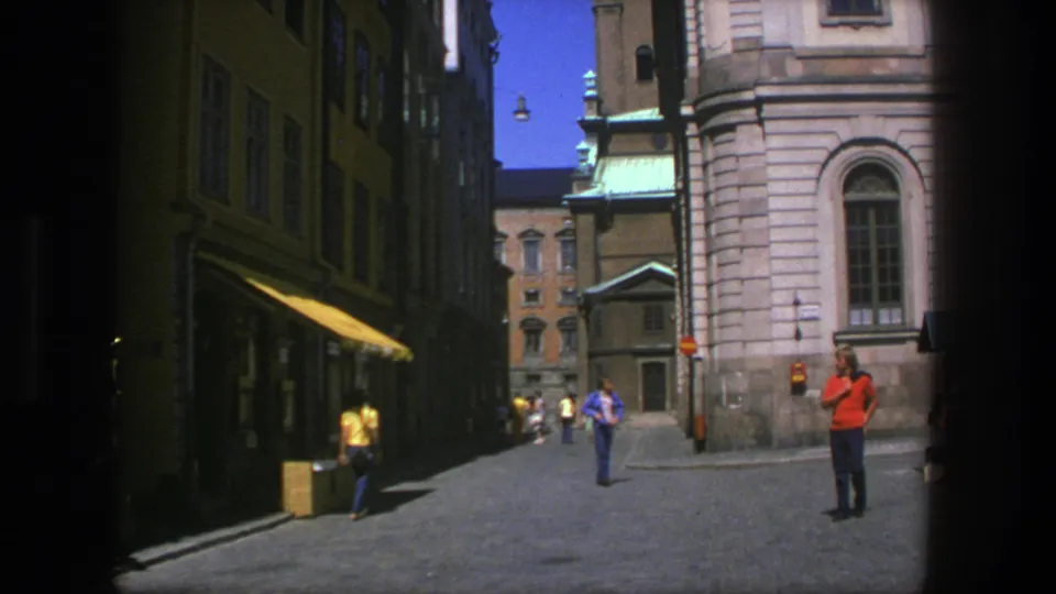 1975 Stockholm alley view with a red-shirted figure, walking pedestrians, shaded storefronts, and a stone church facade in bright daylight.