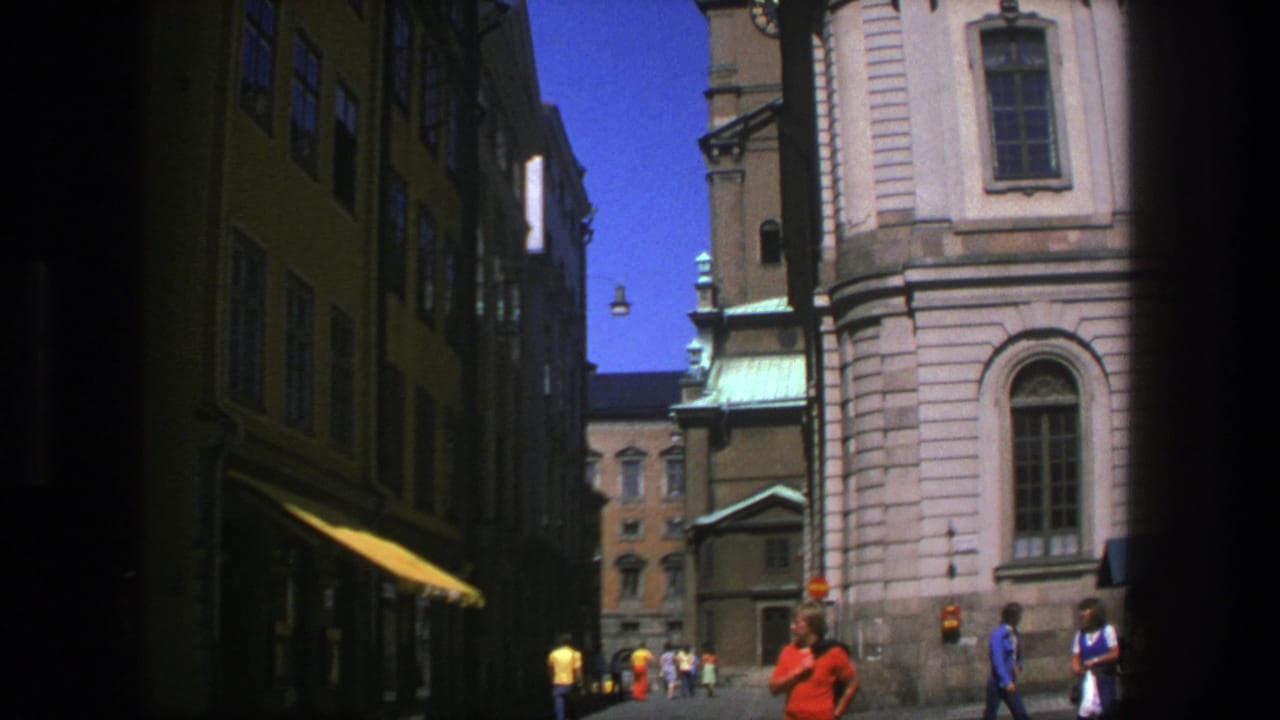 1975 8mm view of pedestrians on a sunlit Stockholm street with shaded facades, hanging lamps, and a pale historic church exterior.