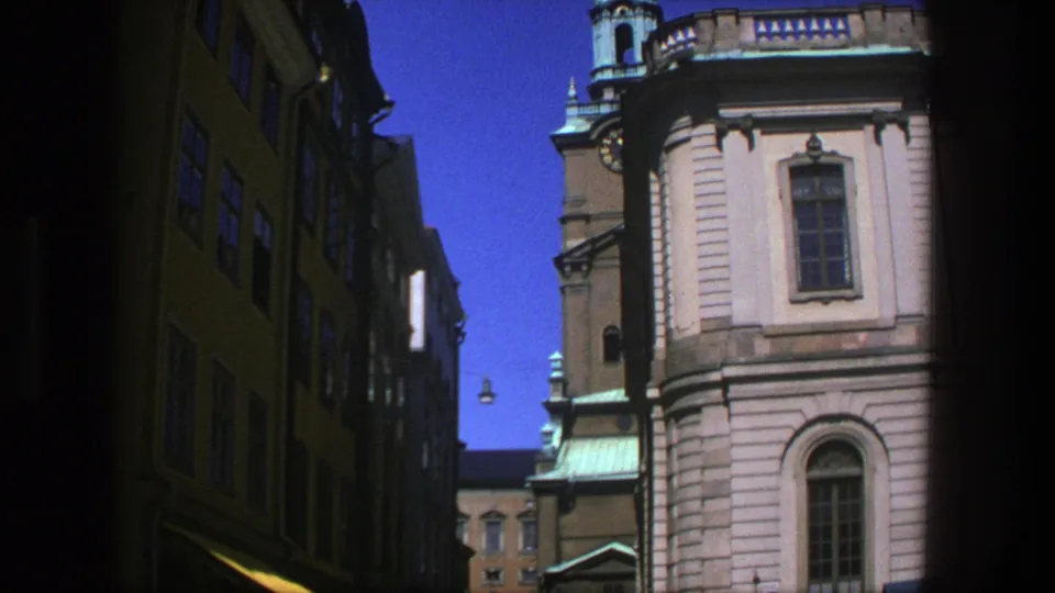 1975 Stockholm 8mm frame looking down a shaded narrow street toward pedestrians and a sunlit historic building with arched windows.