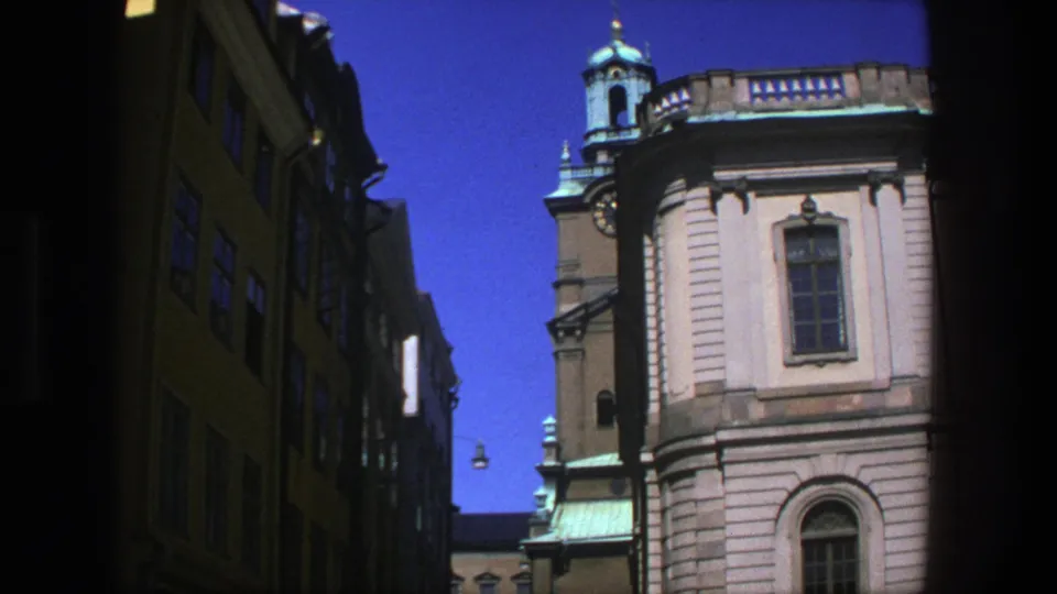 Stockholm 1975 street frame with a pale church-like facade, arched windows, copper roof details, shaded buildings, and pedestrians partly visible.