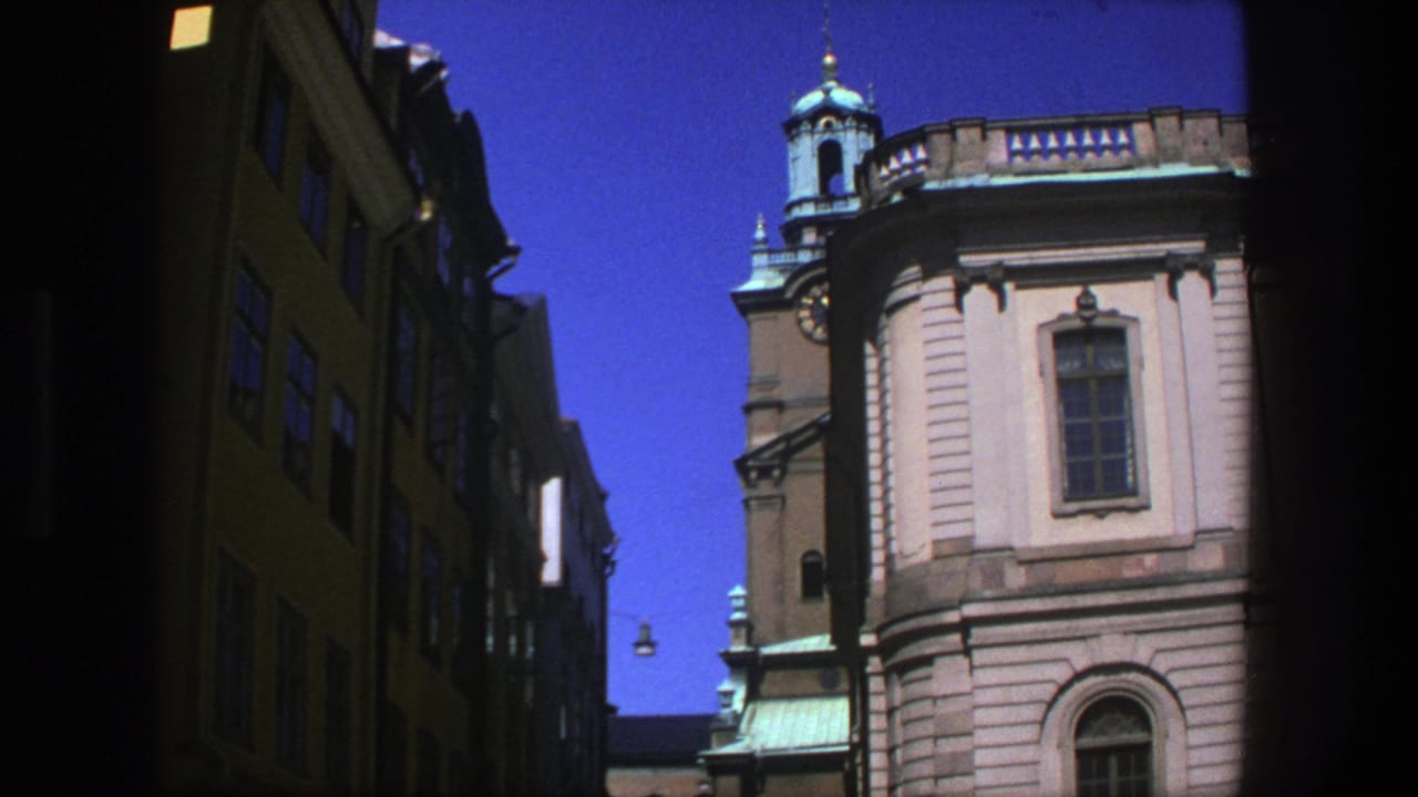 Clock tower and tan facade seen between dark narrow street buildings under blue sky.