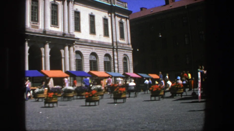Wider 1975 Stockholm square view with colorful awnings, flower carts, pedestrians, cobblestones, and the classical building facade behind.