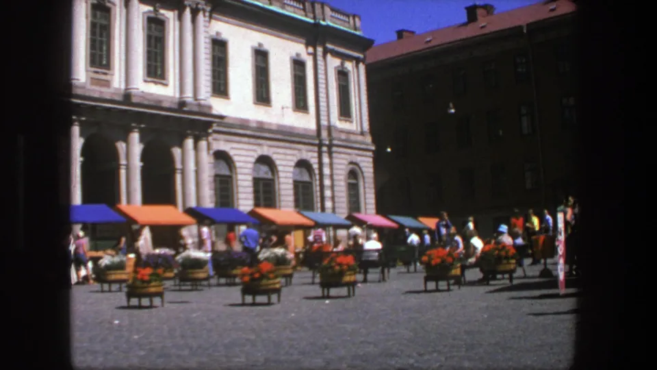 End frame of the 1975 Stockholm square scene, showing pedestrians, bright awnings, flower carts, cobblestones, and historic architecture.