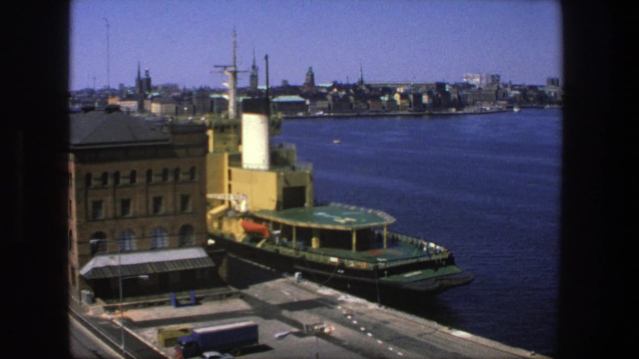 Green boat docked beside a brown waterfront building with blue water and city skyline beyond.