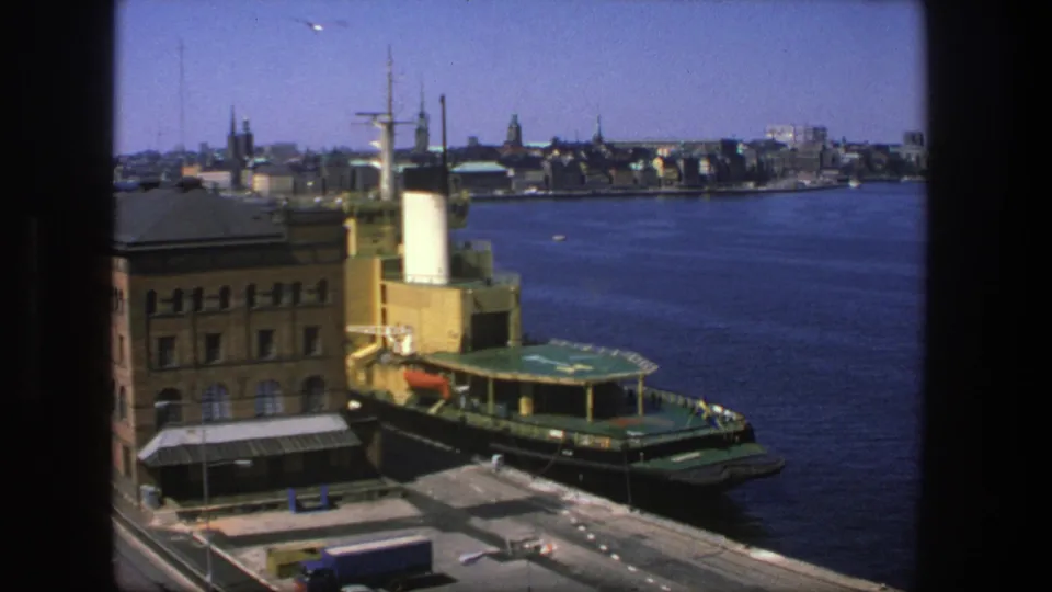 1975 Stockholm harbor frame showing the green boat at the pier with a truck below and skyline spires across the water.