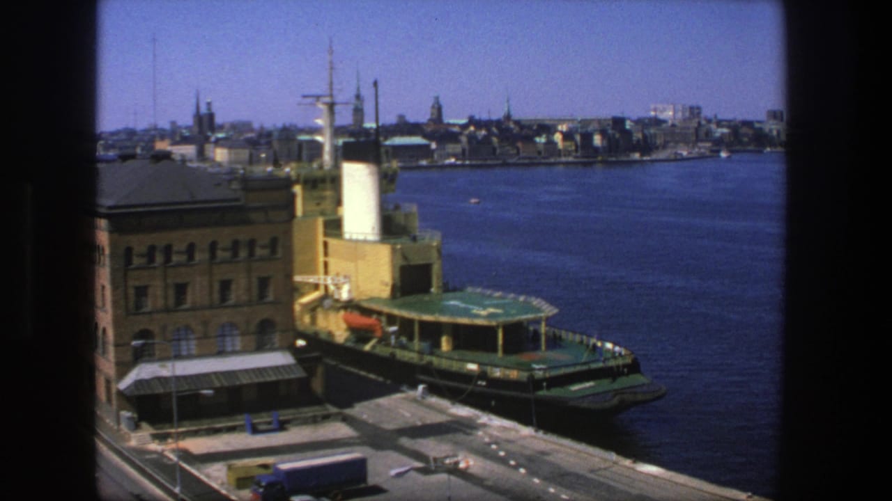 1975 Stockholm harbor view through a dark frame, showing a green ferry at the quay, brick building, blue truck, water, and distant skyline.