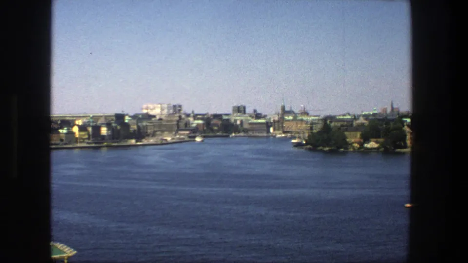 Final wide frame of the 1975 Stockholm harbor scene, showing blue water, distant city buildings, spires, and a small island or shoreline area.