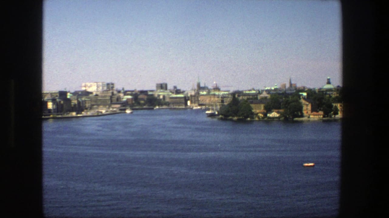 1975 Stockholm waterfront filmed from behind dark window trim, showing blue water, distant urban buildings, green roofs, and a small boat.