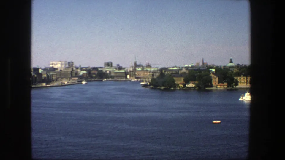 1975 8mm Stockholm view across open water, with a distant shoreline, spires, dark window frame, a small boat, and a larger white vessel.