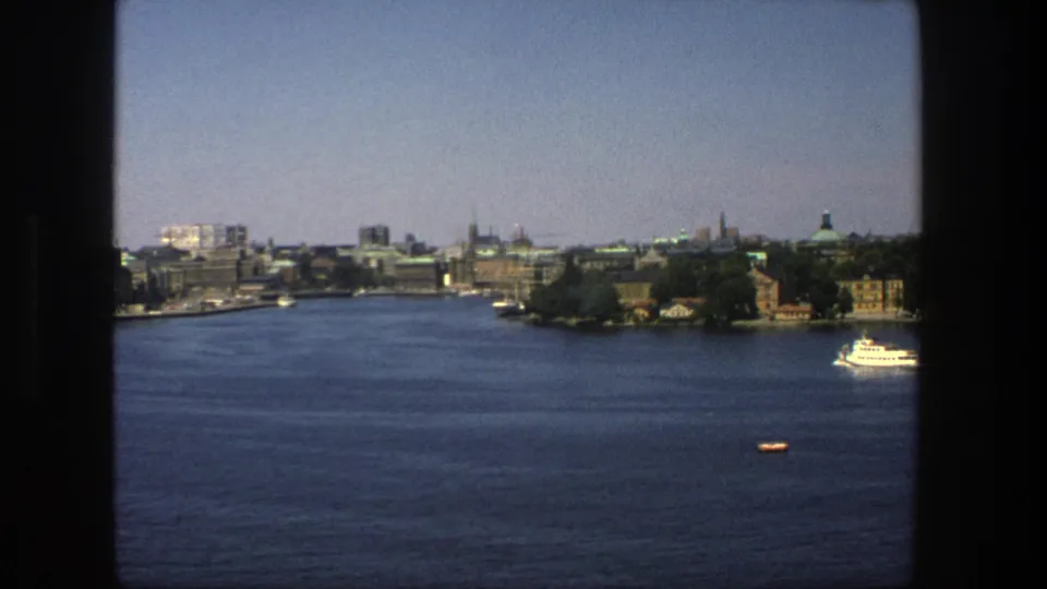 Late-frame 1975 Stockholm waterfront view showing blue water, distant buildings and spires, window trim, a small boat, and a white vessel at right.