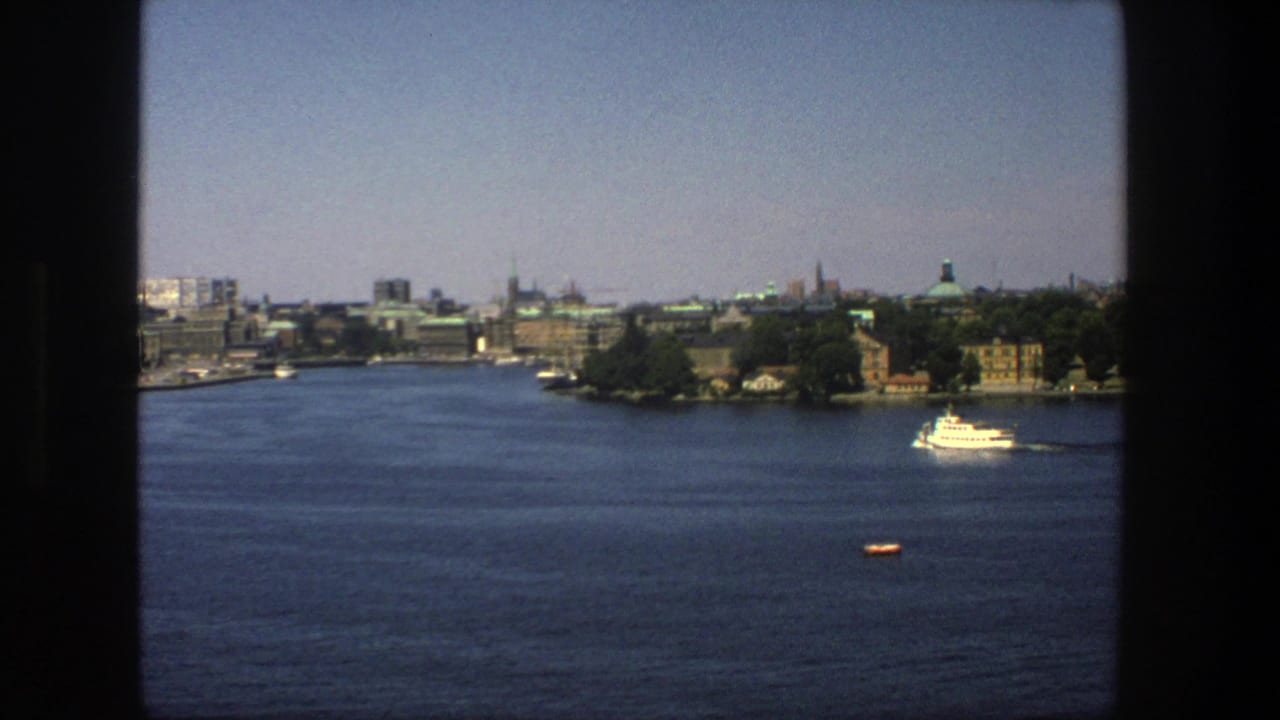 Wide water view with Stockholm skyline, tree-lined shore, and a white boat at right.
