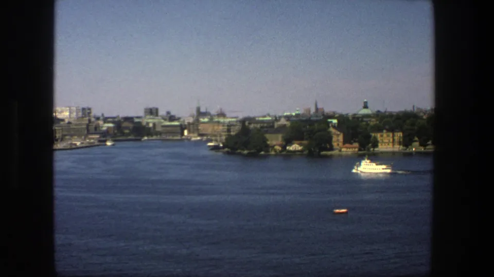 1975 Stockholm skyline across the water, showing a white ferry or tour boat, church spires, waterfront buildings, and dark frame edges.