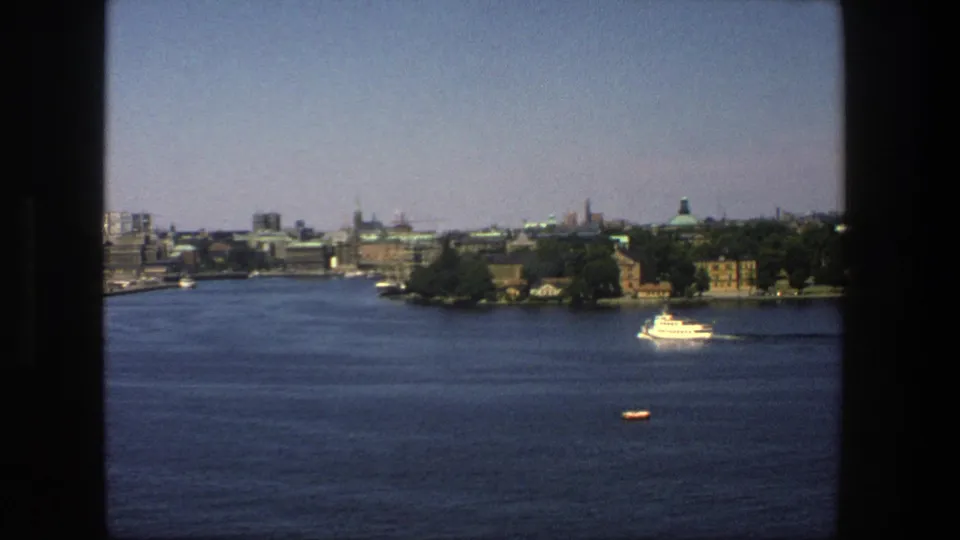1975 home-movie frame of Stockholm's waterfront, with a white boat near the trees, a small orange craft, and distant spires across the water.
