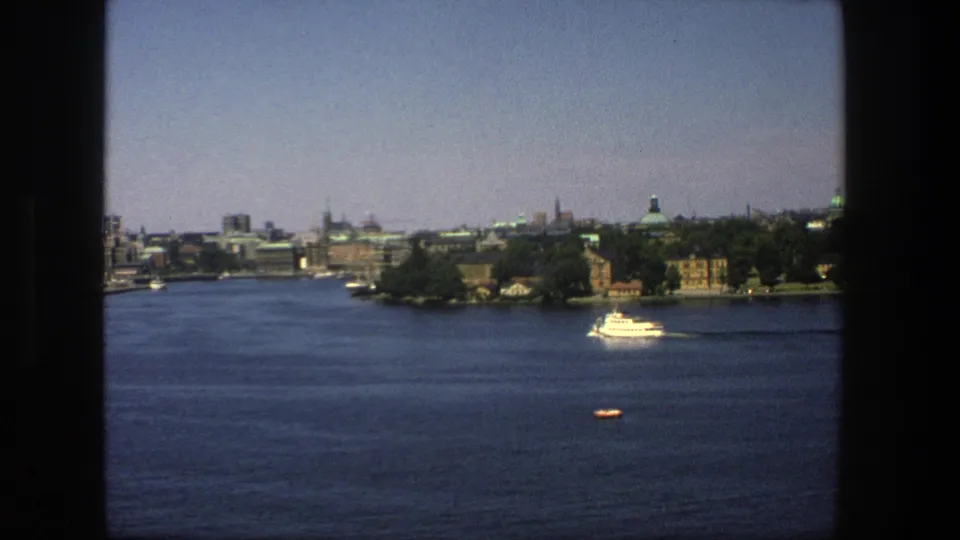 Late-frame 1975 8mm view over Stockholm's water, showing the skyline, tree-lined shore, white boat, and small orange craft within dark window borders.